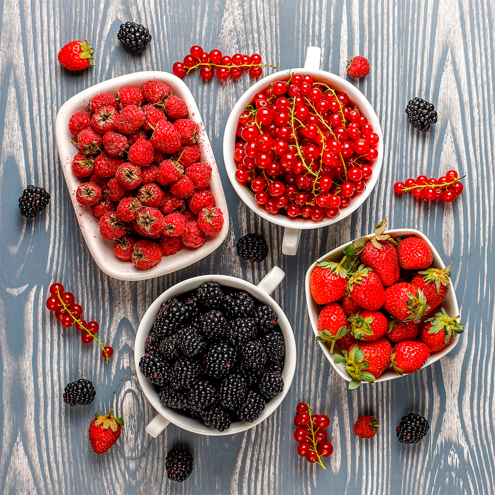Bowls of strawberries, raspberries, blackberries, and red currants on a bluish wooden board.