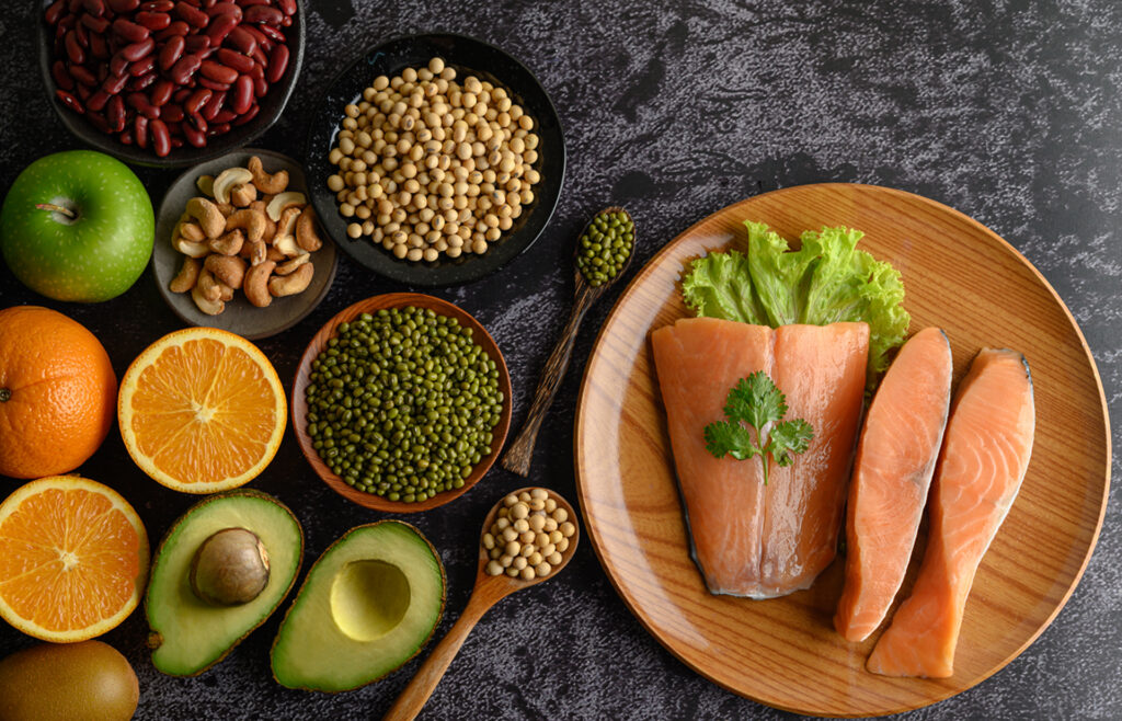 Composition of superfoods on a wooden plate: red beans, soybeans, nuts, salmon, lettuce, oranges, avocado, and kiwi on a textured dark background.