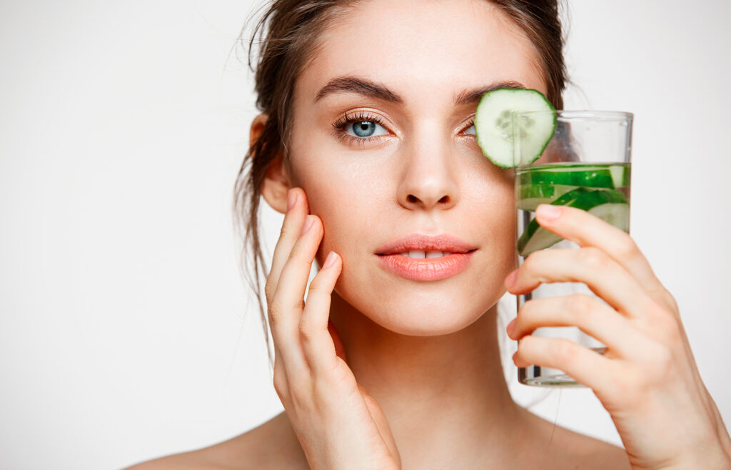 A woman with a cucumber slice over one eye and holding a glass of water with cucumber slices, promoting skin hydration and wellness.