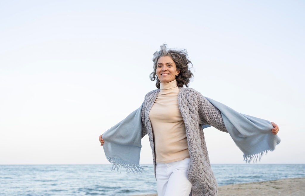 Mature woman with arms open on the beach at sunset, illustrating freedom and well-being in menopause and women's health.