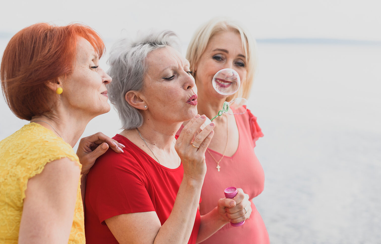 Three mature women laughing and blowing bubbles on the beach, representing joy and connections in the postmenopause phase.