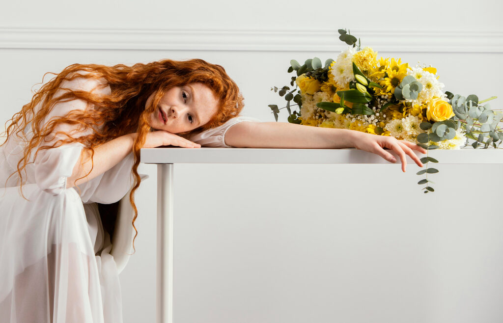 Serene redhead woman with long flowing hair reclining gracefully holding a bouquet of yellow flowers and eucalyptus in a white flowing dress, evoking the gentle guide to feminine intimate health, pleasure, and daily care.